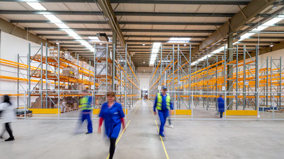 Warehouse workers in high-visibility vests walking along an aisle of tall racks